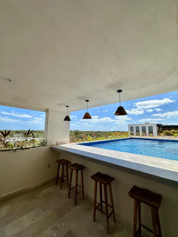 A vibrant rooftop pool bar at TownCenter Zama, featuring a sparkling blue swimming pool, a bar counter with four wooden stools, and modern pendant lights, all set against a clear blue sky with lush green foliage in the background, showcasing a rea...