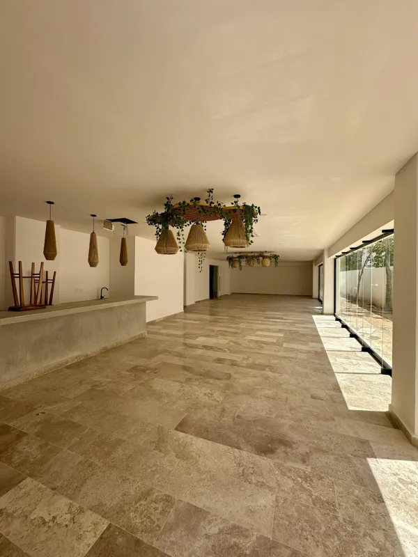 A vibrant rooftop pool bar at TownCenter Zama, featuring a sparkling blue swimming pool, a bar counter with four wooden stools, and modern pendant lights, all set against a clear blue sky with lush green foliage in the background, showcasing a rea...