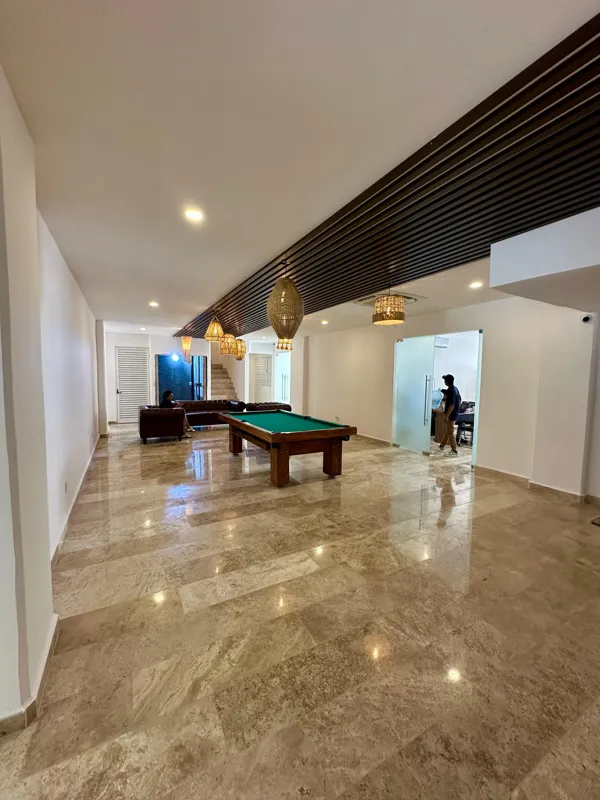 A vibrant rooftop pool bar at TownCenter Zama, featuring a sparkling blue swimming pool, a bar counter with four wooden stools, and modern pendant lights, all set against a clear blue sky with lush green foliage in the background, showcasing a rea...