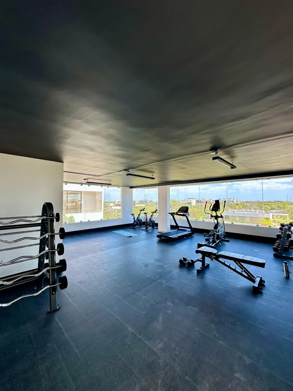 A vibrant rooftop pool bar at TownCenter Zama, featuring a sparkling blue swimming pool, a bar counter with four wooden stools, and modern pendant lights, all set against a clear blue sky with lush green foliage in the background, showcasing a rea...