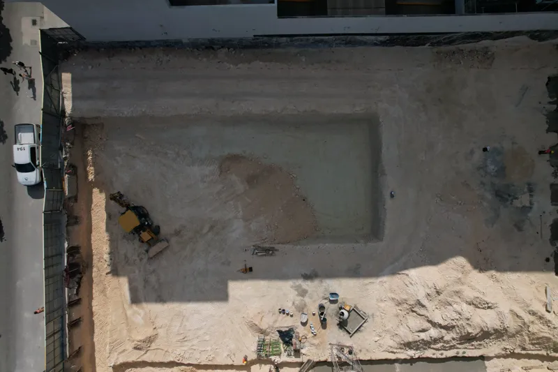 An aerial view of the Solar Midtown development site in Playa del Carmen, showing a large excavation pit for the foundation, with construction equipment visible, surrounded by existing urban buildings and lush greenery, with the Caribbean Sea in t...