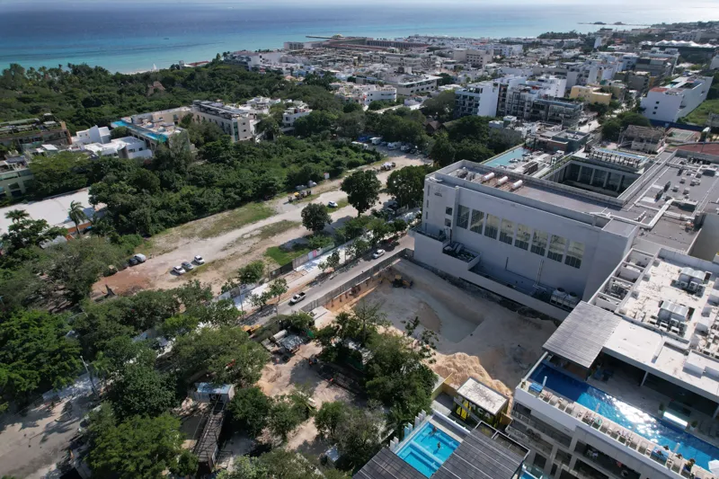 An aerial view of the Solar Midtown development site in Playa del Carmen, showing a large excavation pit for the foundation, with construction equipment visible, surrounded by existing urban buildings and lush greenery, with the Caribbean Sea in t...