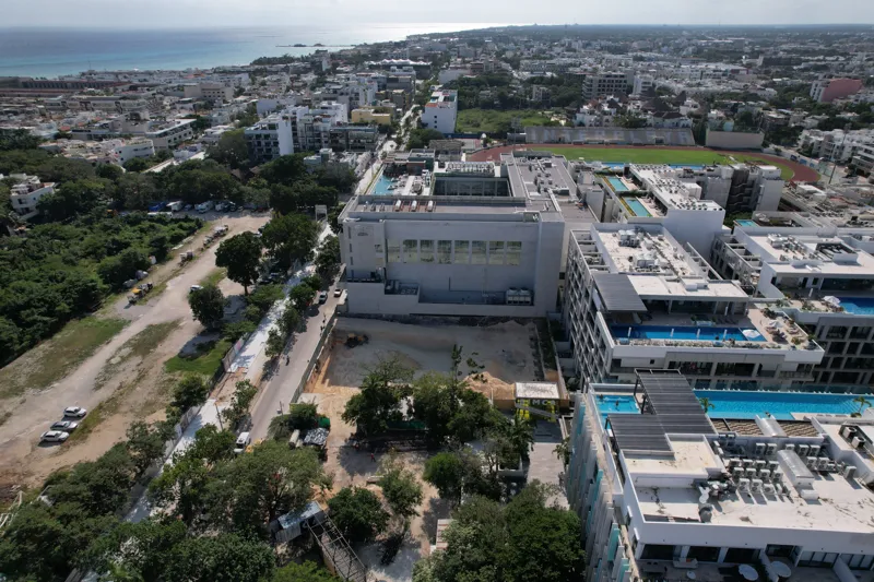 An aerial view of the Solar Midtown development site in Playa del Carmen, showing a large excavation pit for the foundation, with construction equipment visible, surrounded by existing urban buildings and lush greenery, with the Caribbean Sea in t...