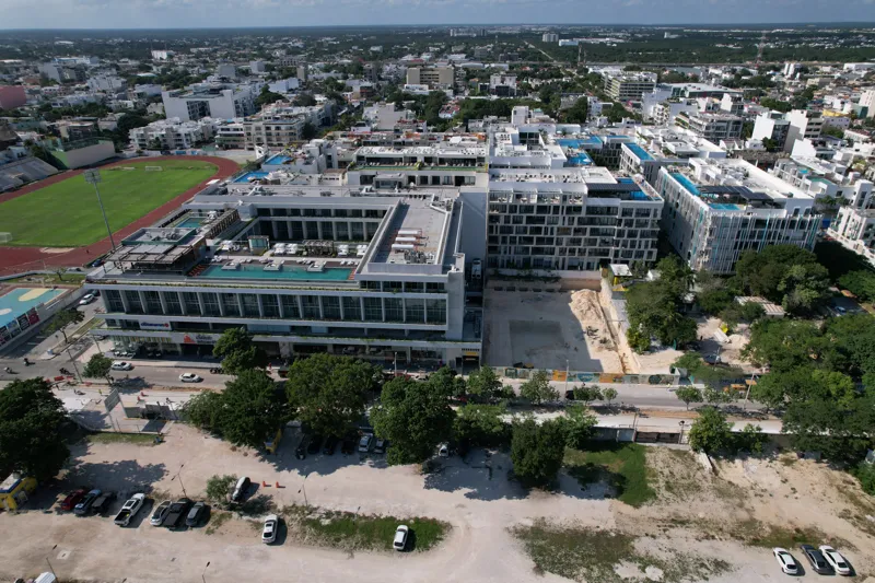 An aerial view of the Solar Midtown development site in Playa del Carmen, showing a large excavation pit for the foundation, with construction equipment visible, surrounded by existing urban buildings and lush greenery, with the Caribbean Sea in t...