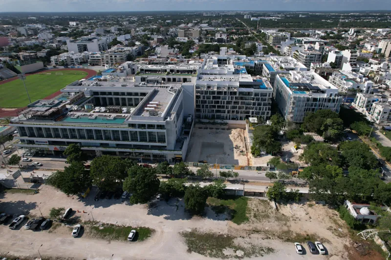An aerial view of the Solar Midtown development site in Playa del Carmen, showing a large excavation pit for the foundation, with construction equipment visible, surrounded by existing urban buildings and lush greenery, with the Caribbean Sea in t...