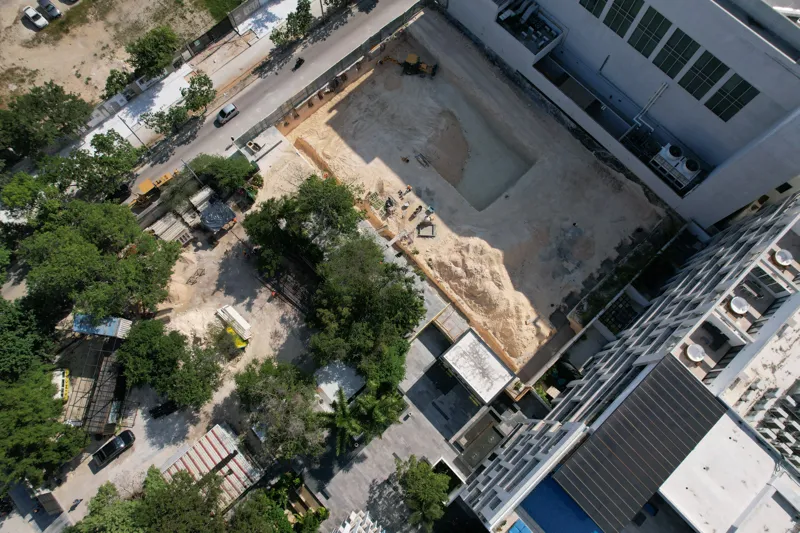 An aerial view of the Solar Midtown development site in Playa del Carmen, showing a large excavation pit for the foundation, with construction equipment visible, surrounded by existing urban buildings and lush greenery, with the Caribbean Sea in t...