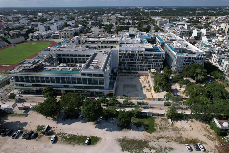 An aerial view of the Solar Midtown development site in Playa del Carmen, showing a large excavation pit for the foundation, with construction equipment visible, surrounded by existing urban buildings and lush greenery, with the Caribbean Sea in t...