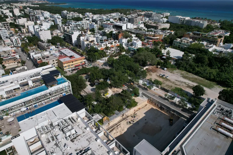 An aerial view of the Solar Midtown development site in Playa del Carmen, showing a large excavation pit for the foundation, with construction equipment visible, surrounded by existing urban buildings and lush greenery, with the Caribbean Sea in t...