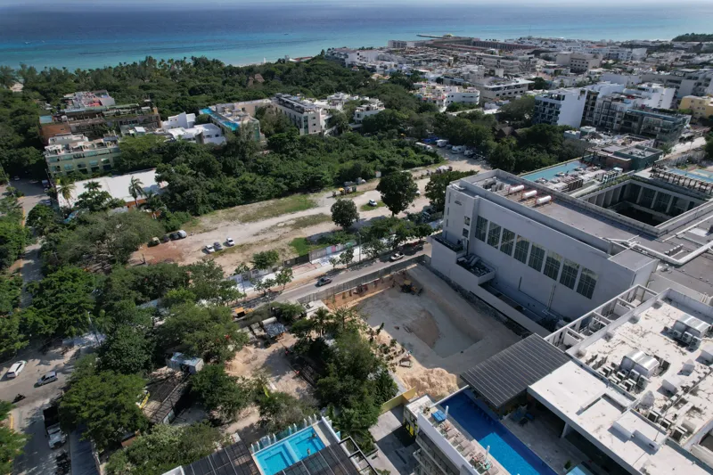 An aerial view of the Solar Midtown development site in Playa del Carmen, showing a large excavation pit for the foundation, with construction equipment visible, surrounded by existing urban buildings and lush greenery, with the Caribbean Sea in t...