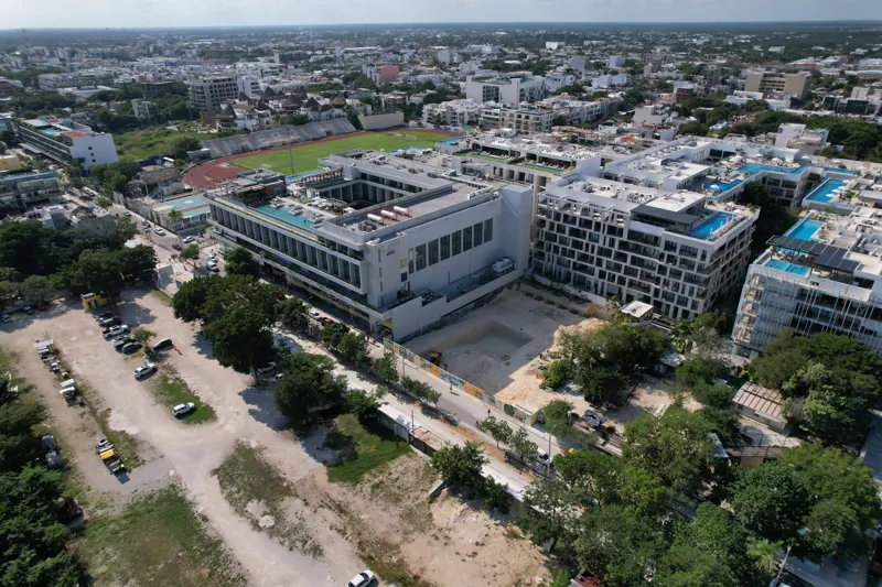 An aerial view of the Solar Midtown development site in Playa del Carmen, showing a large excavation pit for the foundation, with construction equipment visible, surrounded by existing urban buildings and lush greenery, with the Caribbean Sea in t...