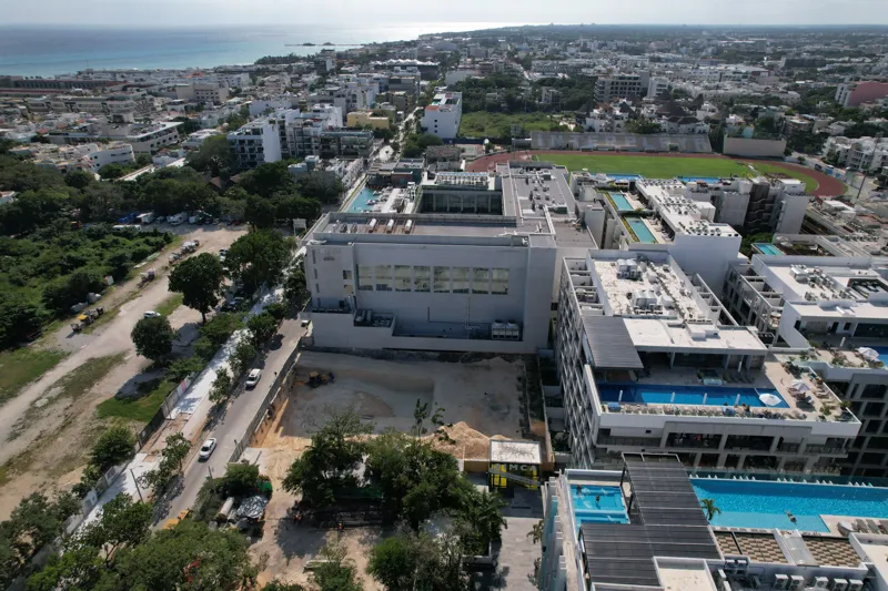 An aerial view of the Solar Midtown development site in Playa del Carmen, showing a large excavation pit for the foundation, with construction equipment visible, surrounded by existing urban buildings and lush greenery, with the Caribbean Sea in t...