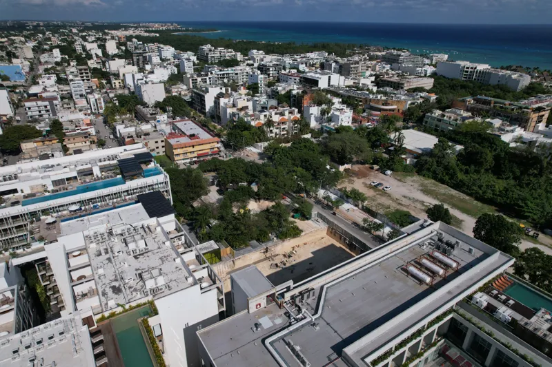 An aerial view of the Solar Midtown development site in Playa del Carmen, showing a large excavation pit for the foundation, with construction equipment visible, surrounded by existing urban buildings and lush greenery, with the Caribbean Sea in t...
