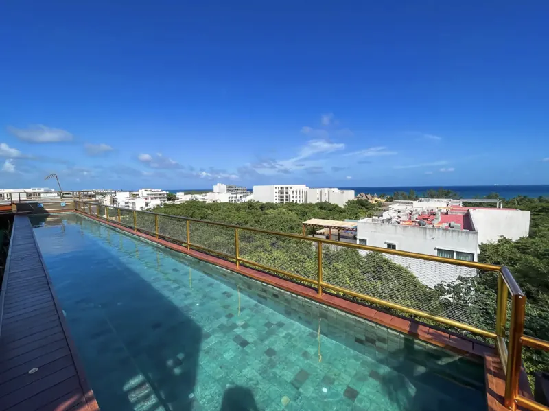 Aerial view of Residencia Condos in Playa del Carmen, featuring a large, completed rooftop infinity pool with people swimming, surrounded by lounge areas and lush greenery, with the Caribbean Sea visible in the background under a partly cloudy sky.