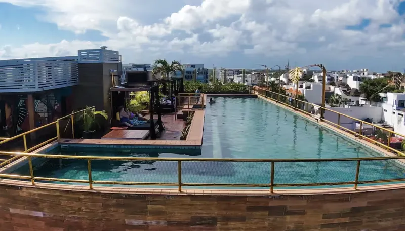 Aerial view of Residencia Condos in Playa del Carmen, featuring a large, completed rooftop infinity pool with people swimming, surrounded by lounge areas and lush greenery, with the Caribbean Sea visible in the background under a partly cloudy sky.