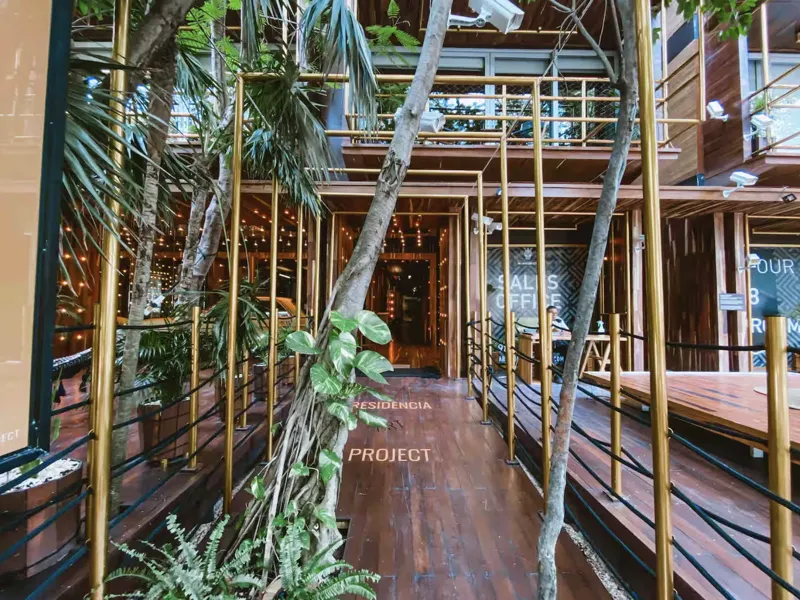 Aerial view of Residencia Condos in Playa del Carmen, featuring a large, completed rooftop infinity pool with people swimming, surrounded by lounge areas and lush greenery, with the Caribbean Sea visible in the background under a partly cloudy sky.