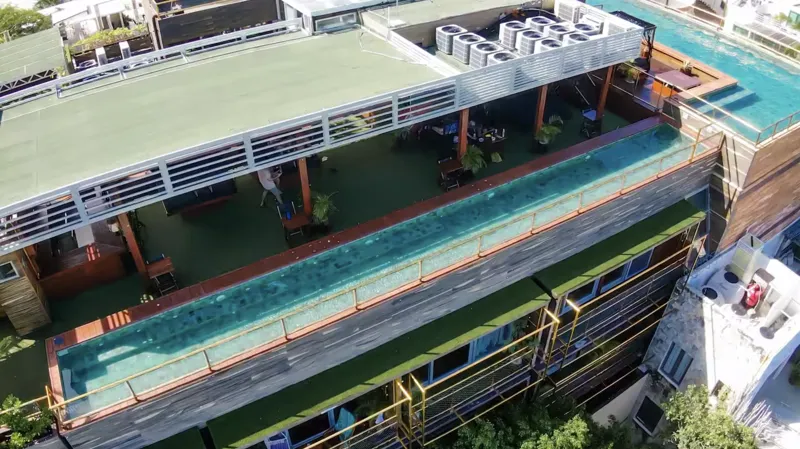 Aerial view of Residencia Condos in Playa del Carmen, featuring a large, completed rooftop infinity pool with people swimming, surrounded by lounge areas and lush greenery, with the Caribbean Sea visible in the background under a partly cloudy sky.