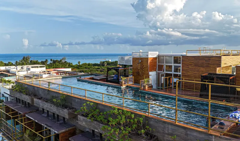 Aerial view of Residencia Condos in Playa del Carmen, featuring a large, completed rooftop infinity pool with people swimming, surrounded by lounge areas and lush greenery, with the Caribbean Sea visible in the background under a partly cloudy sky.