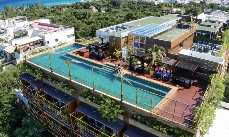 Aerial view of Residencia Condos in Playa del Carmen, featuring a large, completed rooftop infinity pool with people swimming, surrounded by lounge areas and lush greenery, with the Caribbean Sea visible in the background under a partly cloudy sky.