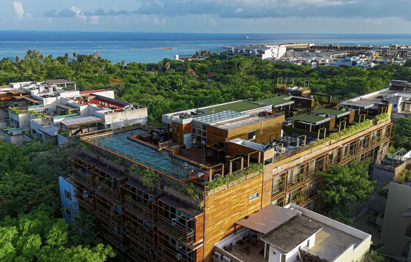Aerial view of Residencia Condos in Playa del Carmen, featuring a large, completed rooftop infinity pool with people swimming, surrounded by lounge areas and lush greenery, with the Caribbean Sea visible in the background under a partly cloudy sky.