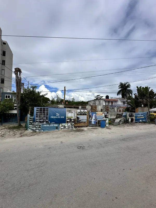 An active construction site for the Piedra de Mar development under a bright blue sky, featuring concrete mixers, extensive scaffolding, rebar, and multiple workers engaged in building structural elements, with a crane visible in the background.
