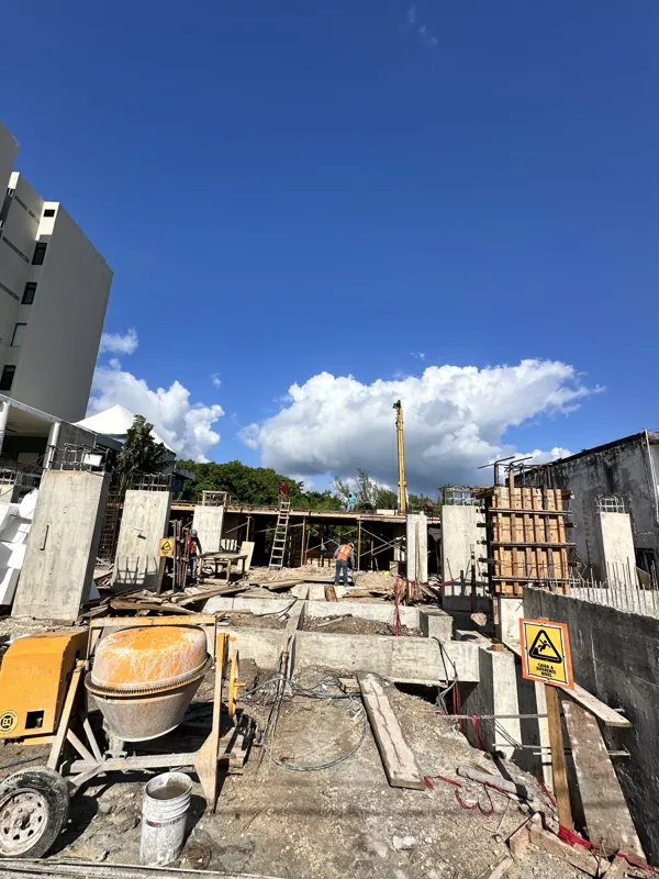 An active construction site for the Piedra de Mar development under a bright blue sky, featuring concrete mixers, extensive scaffolding, rebar, and multiple workers engaged in building structural elements, with a crane visible in the background.