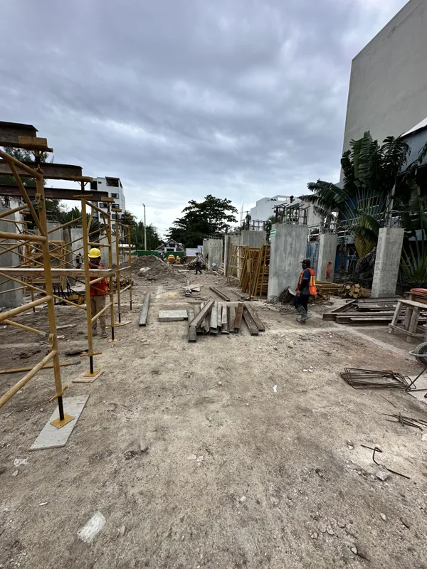 An active construction site for the Piedra de Mar development under a bright blue sky, featuring concrete mixers, extensive scaffolding, rebar, and multiple workers engaged in building structural elements, with a crane visible in the background.