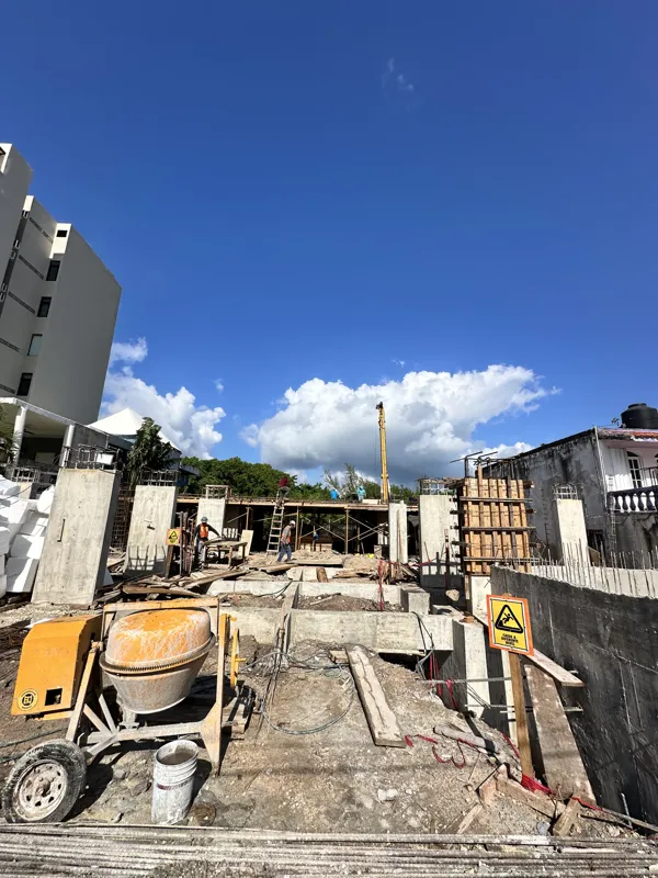 An active construction site for the Piedra de Mar development under a bright blue sky, featuring concrete mixers, extensive scaffolding, rebar, and multiple workers engaged in building structural elements, with a crane visible in the background.