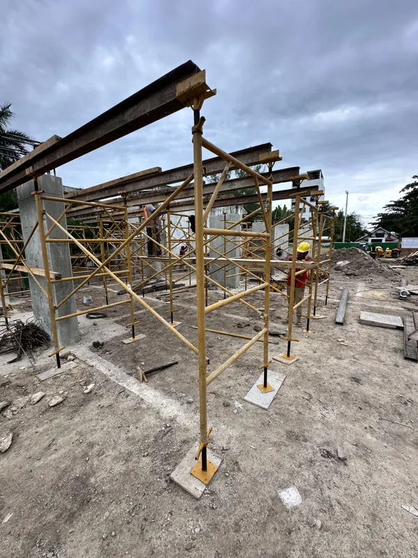 An active construction site for the Piedra de Mar development under a bright blue sky, featuring concrete mixers, extensive scaffolding, rebar, and multiple workers engaged in building structural elements, with a crane visible in the background.