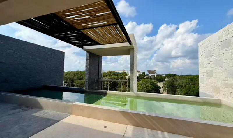 Exterior view of the Xexen y Xakah development showing significant construction progress, with scaffolding in front of the main entrance, stone cladding on the facade, and a pickup truck parked on the gravel driveway under a clear blue sky.