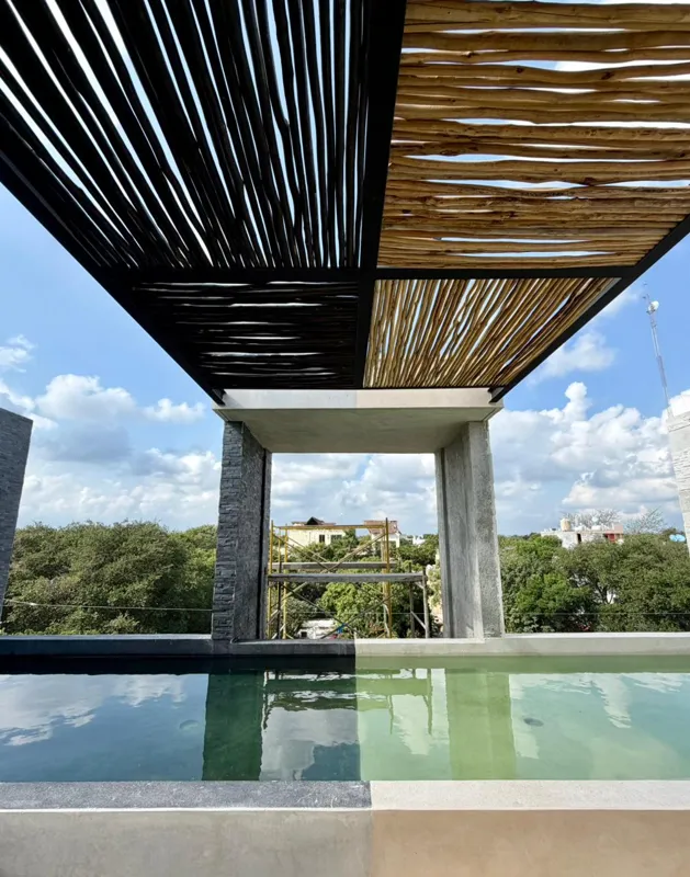 Exterior view of the Xexen y Xakah development showing significant construction progress, with scaffolding in front of the main entrance, stone cladding on the facade, and a pickup truck parked on the gravel driveway under a clear blue sky.