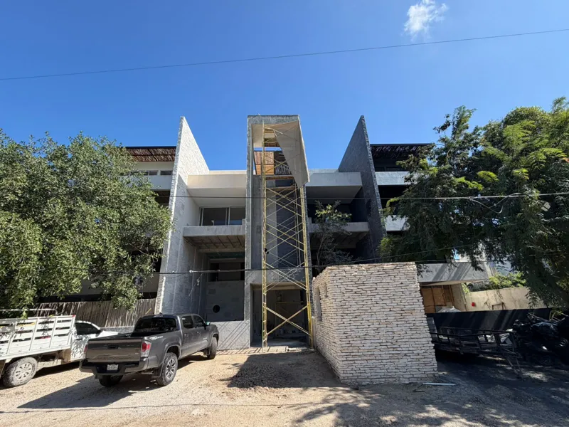 Exterior view of the Xexen y Xakah development showing significant construction progress, with scaffolding in front of the main entrance, stone cladding on the facade, and a pickup truck parked on the gravel driveway under a clear blue sky.