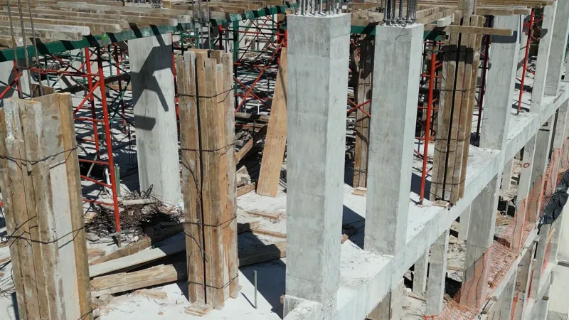 An overhead view of the Maia development construction site, showing multiple grey concrete columns and horizontal beams forming the building's structure. Wooden formwork is still visible around some columns, secured with wire, and red and green sc...