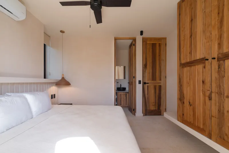 A bright, sunny rooftop terrace featuring a private swimming pool with clear blue water, surrounded by lush green plants and a light-colored building facade. A shaded pergola covers an outdoor counter area with wooden cabinets, all under a clear b...