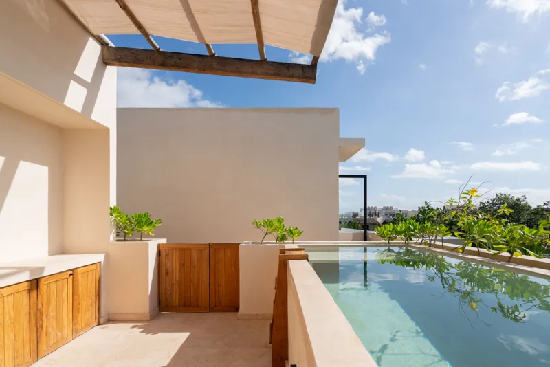 A bright, sunny rooftop terrace featuring a private swimming pool with clear blue water, surrounded by lush green plants and a light-colored building facade. A shaded pergola covers an outdoor counter area with wooden cabinets, all under a clear b...