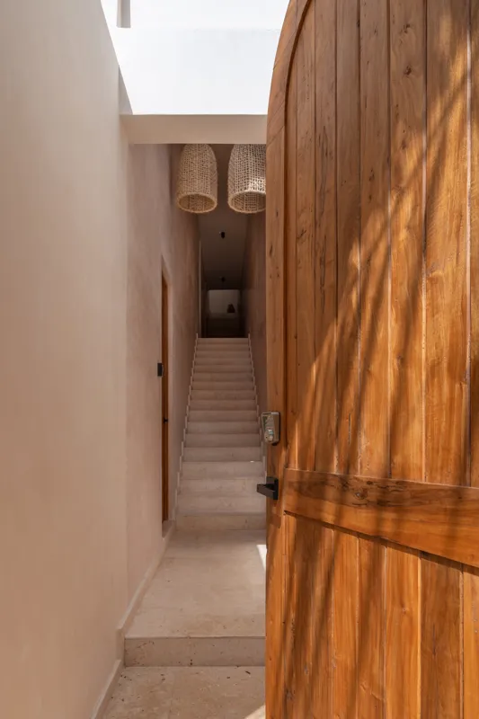 A bright, sunny rooftop terrace featuring a private swimming pool with clear blue water, surrounded by lush green plants and a light-colored building facade. A shaded pergola covers an outdoor counter area with wooden cabinets, all under a clear b...