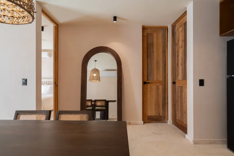 A bright, sunny rooftop terrace featuring a private swimming pool with clear blue water, surrounded by lush green plants and a light-colored building facade. A shaded pergola covers an outdoor counter area with wooden cabinets, all under a clear b...