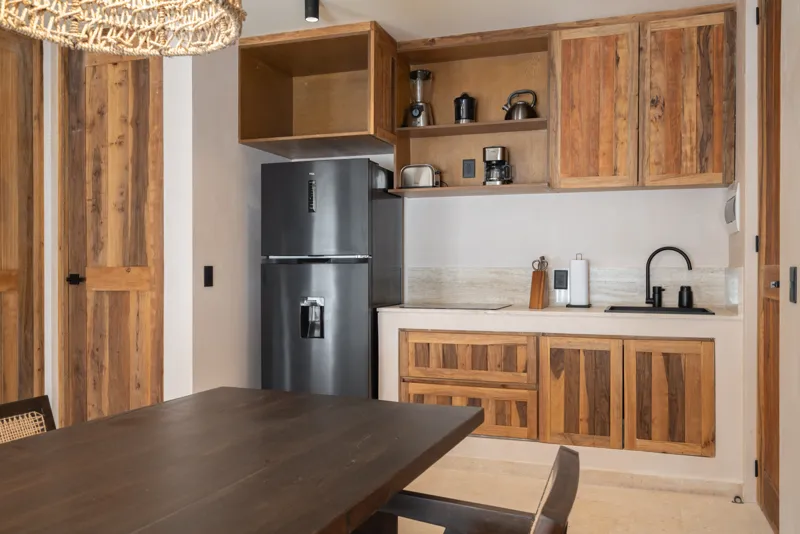 A bright, sunny rooftop terrace featuring a private swimming pool with clear blue water, surrounded by lush green plants and a light-colored building facade. A shaded pergola covers an outdoor counter area with wooden cabinets, all under a clear b...