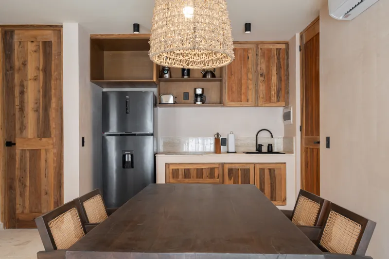 A bright, sunny rooftop terrace featuring a private swimming pool with clear blue water, surrounded by lush green plants and a light-colored building facade. A shaded pergola covers an outdoor counter area with wooden cabinets, all under a clear b...