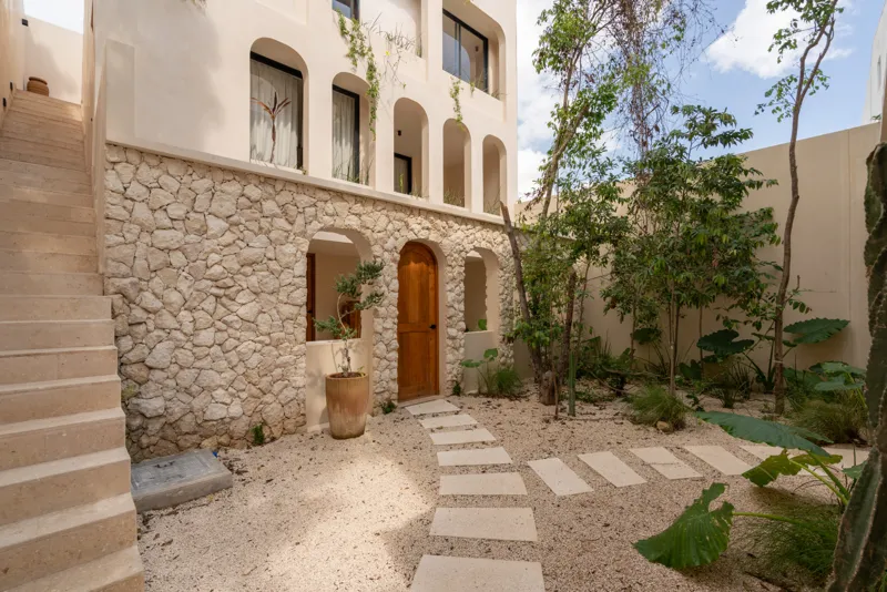 A bright, sunny rooftop terrace featuring a private swimming pool with clear blue water, surrounded by lush green plants and a light-colored building facade. A shaded pergola covers an outdoor counter area with wooden cabinets, all under a clear b...