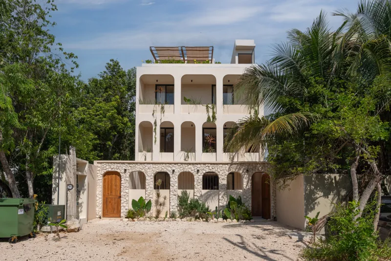 A bright, sunny rooftop terrace featuring a private swimming pool with clear blue water, surrounded by lush green plants and a light-colored building facade. A shaded pergola covers an outdoor counter area with wooden cabinets, all under a clear b...