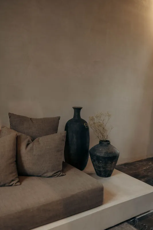 An expansive outdoor lounge area at La Reserva Tulum, featuring dark walls, built-in seating with plush cushions, natural tree trunks integrated into the design, woven pendant lights, large decorative vases, and a green patterned rug, all set agai...