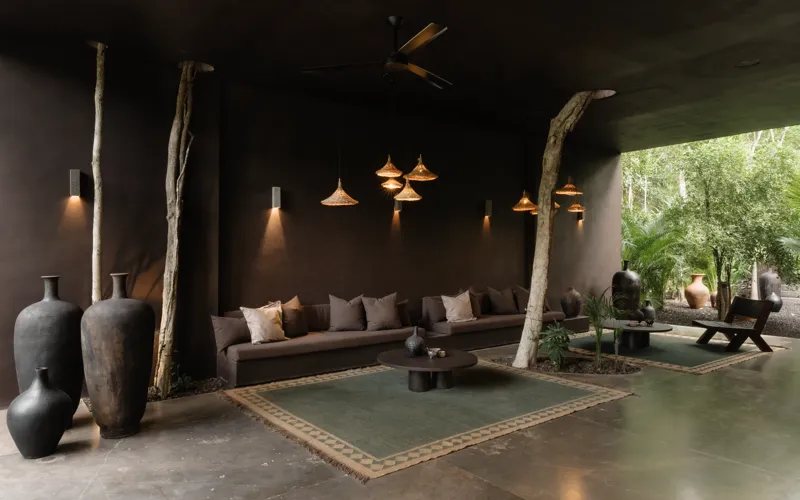 An expansive outdoor lounge area at La Reserva Tulum, featuring dark walls, built-in seating with plush cushions, natural tree trunks integrated into the design, woven pendant lights, large decorative vases, and a green patterned rug, all set agai...