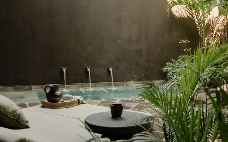 An expansive outdoor lounge area at La Reserva Tulum, featuring dark walls, built-in seating with plush cushions, natural tree trunks integrated into the design, woven pendant lights, large decorative vases, and a green patterned rug, all set agai...