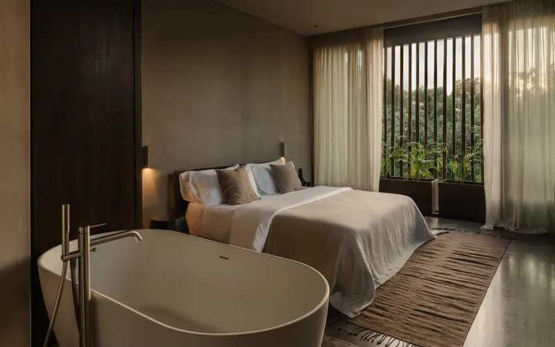 An expansive outdoor lounge area at La Reserva Tulum, featuring dark walls, built-in seating with plush cushions, natural tree trunks integrated into the design, woven pendant lights, large decorative vases, and a green patterned rug, all set agai...