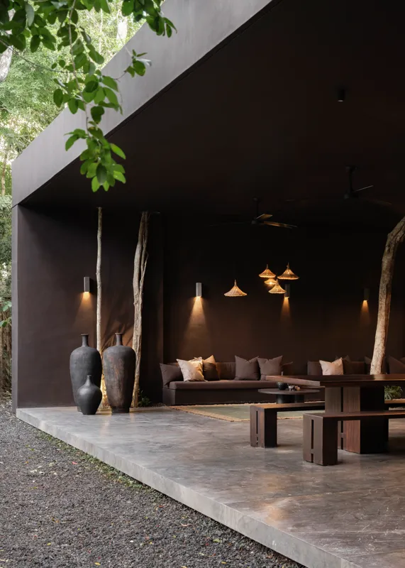 An expansive outdoor lounge area at La Reserva Tulum, featuring dark walls, built-in seating with plush cushions, natural tree trunks integrated into the design, woven pendant lights, large decorative vases, and a green patterned rug, all set agai...