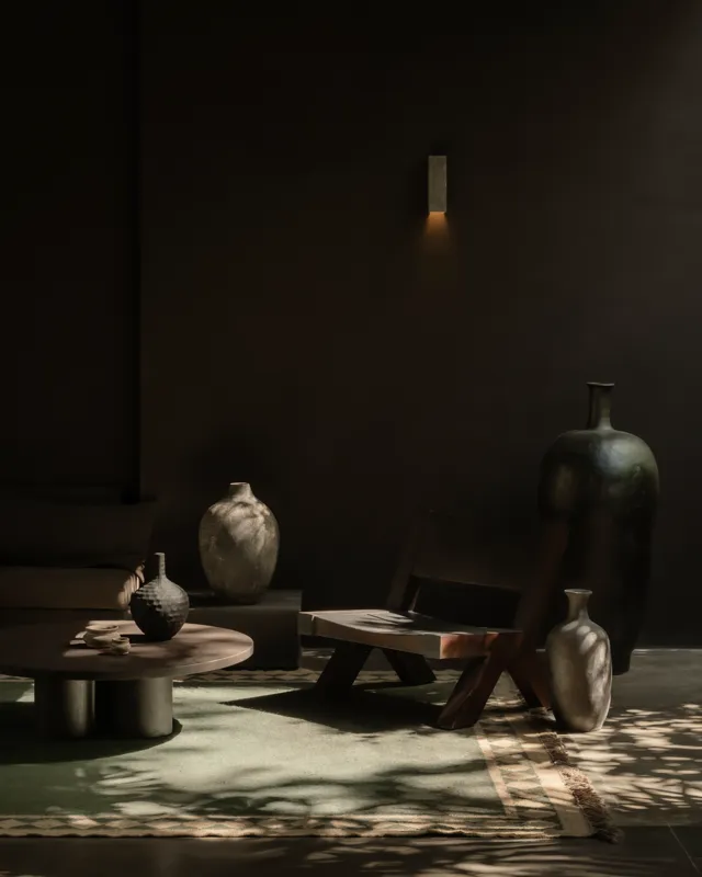 An expansive outdoor lounge area at La Reserva Tulum, featuring dark walls, built-in seating with plush cushions, natural tree trunks integrated into the design, woven pendant lights, large decorative vases, and a green patterned rug, all set agai...