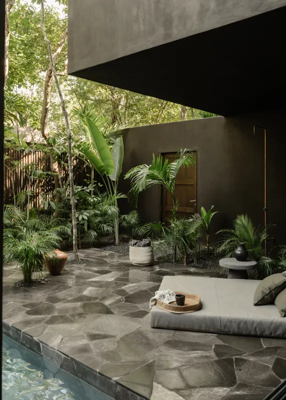 An expansive outdoor lounge area at La Reserva Tulum, featuring dark walls, built-in seating with plush cushions, natural tree trunks integrated into the design, woven pendant lights, large decorative vases, and a green patterned rug, all set agai...