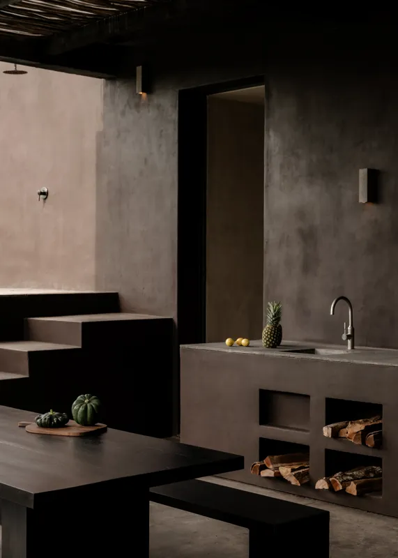 An expansive outdoor lounge area at La Reserva Tulum, featuring dark walls, built-in seating with plush cushions, natural tree trunks integrated into the design, woven pendant lights, large decorative vases, and a green patterned rug, all set agai...