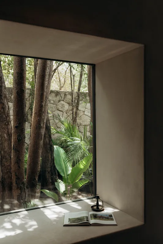 An expansive outdoor lounge area at La Reserva Tulum, featuring dark walls, built-in seating with plush cushions, natural tree trunks integrated into the design, woven pendant lights, large decorative vases, and a green patterned rug, all set agai...