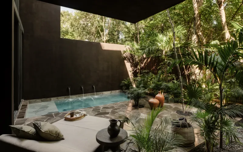 An expansive outdoor lounge area at La Reserva Tulum, featuring dark walls, built-in seating with plush cushions, natural tree trunks integrated into the design, woven pendant lights, large decorative vases, and a green patterned rug, all set agai...